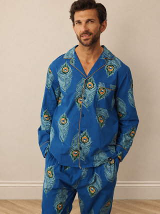 A man wearing a two-piece pyjama set with a vibrant blue base and a detailed peacock feather print, standing against a neutral beige wall.