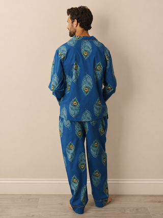 A man stands with his back to the camera wearing a matching two-piece pyjama set in a vibrant blue colour, featuring a detailed peacock feather print in shades of teal, green, and gold. The set includes a long-sleeve top and matching pants, photographed against a neutral beige wall and light wood floor.