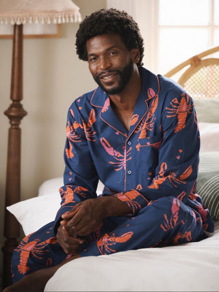 Male model wears a pair of Navy pyjamas with lobster print sat on a bed. 