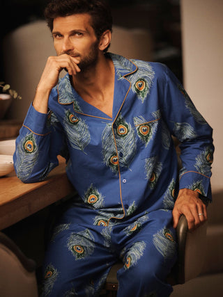 A man wearing a blue pyjama set with a peacock feather print and gold piping, sitting in a chair in a warm, dimly lit indoor setting.