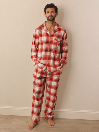 A man wearing a red and white plaid pyjama set stands barefoot against a plain beige wall on a light wood floor.