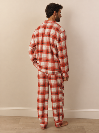 A man stands with his back to the camera wearing a two-piece long-sleeve pyjama set in a red, orange, and white plaid pattern, set against a plain beige wall and light wood floor.