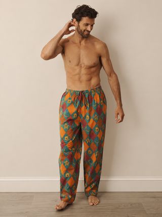 A shirtless man wearing patterned lounge pyjama pants with an orange, teal, and brown geometric design, standing against a plain beige wall.