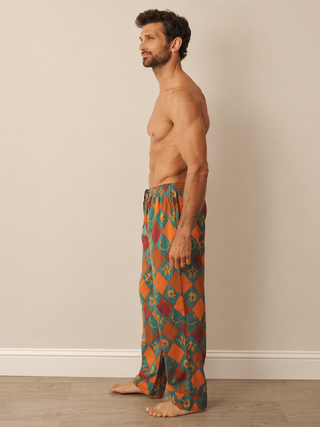 A man stands in profile wearing loose, patterned lounge pants with an orange, teal, and red geometric design, photographed indoors against a plain beige wall and light wood floor.