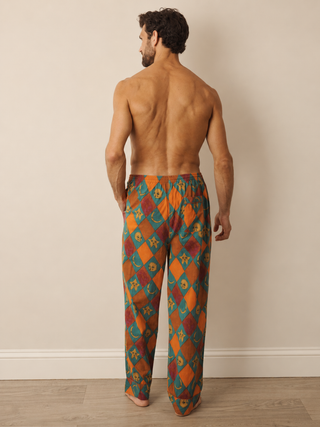 A man stands with his back to the camera wearing loose, wide-leg pyjama pants with a colourful harlequin diamond pattern in orange, teal, red, and yellow, set against a neutral beige wall and light wood floor.