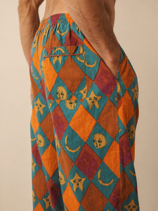 A close-up of a person wearing loose patterned pyjama bottoms with a colourful harlequin diamond print in orange, teal, purple, and mustard yellow, featuring moon and star motifs.