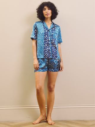 A woman modelling a two-piece pyjama set consisting of a short-sleeve button-up shirt and matching shorts, both in a blue base with a white abstract spotted pattern, standing against a beige wall.