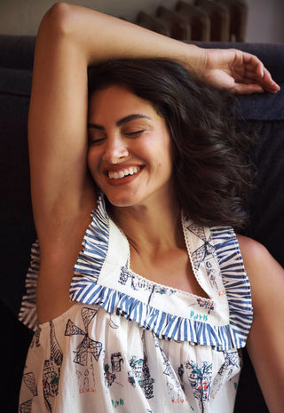 A woman smiling with her arm raised, wearing a sleeveless white nightdress with blue striped ruffle trim and a multi-colour sketch print.