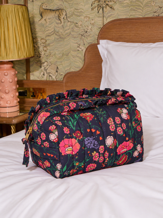 A quilted cosmetic pouch with a dark navy floral print sits on a white bed in a bedroom setting, next to a pink lamp and wooden headboard.