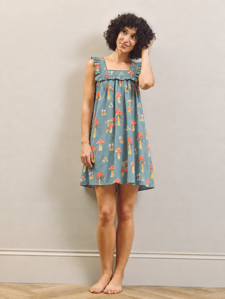 A woman stands barefoot against a plain light gray wall wearing a sleeveless blue-gray nightgown with a red, orange, and yellow mushroom print.