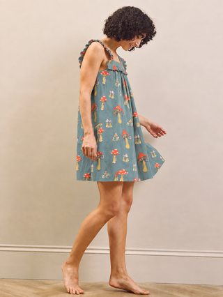 A woman wearing a sleeveless, ruffled-shoulder babydoll nightgown with a blue-green background and a red and yellow mushroom print, standing barefoot against a neutral beige wall.
