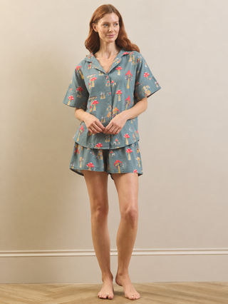 A woman stands barefoot in a bedroom setting wearing a two-piece pyjama set. The set consists of a short-sleeve button-up shirt and matching shorts in a muted blue-gray color, both printed with a whimsical pattern of red mushrooms, yellow flowers, and green leaves.