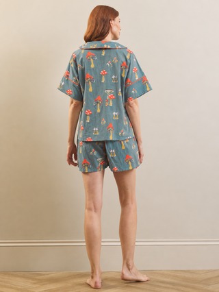 A woman stands barefoot against a beige wall wearing a matching pyjama set with a blue-gray base and a colourful mushroom print, shown from the back.