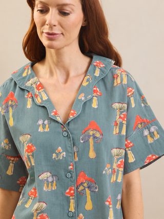A woman wearing a short-sleeve button-front pyjama shirt in muted blue-green with an allover mushroom print in red, yellow, and gray, photographed against a plain beige studio background.