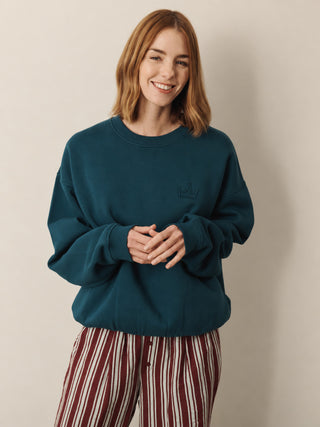 A woman with shoulder-length auburn hair smiling, wearing a dark teal crewneck sweatshirt with a small black crown logo on the left chest, paired with red and white vertically striped pants, against a plain beige studio background.