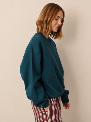 A woman with shoulder-length auburn hair is shown in profile wearing an oversized dark teal crewneck sweatshirt and red-and-white vertically striped pants, set against a plain beige studio background.