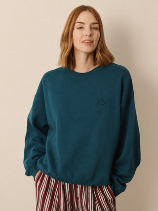 A woman with shoulder-length auburn hair wearing a dark teal crewneck sweatshirt with a small black embroidered crown on the left chest, paired with red and white striped pants, against a solid beige background.