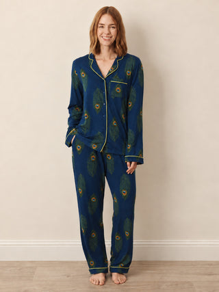 A woman modelling a two-piece pyjama set featuring a dark teal blue base with a green and gold peacock feather pattern, accented with gold piping on a beige background.
