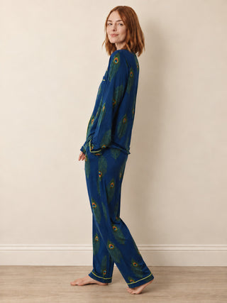 A woman modelling a two-piece pyjama set with a blue base and green peacock feather print, standing barefoot against a beige wall.