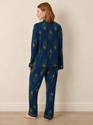 A woman stands barefoot against a plain beige wall, wearing a matching long-sleeve pyjama set with a dark blue base and an allover peacock feather print. The set is shown from the back, highlighting the collar, cuffs, and the pattern details.