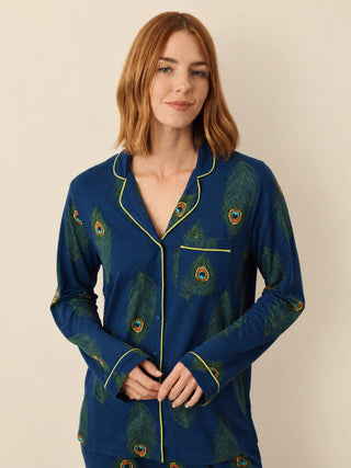 A woman wearing a long-sleeve pyjama top with a peacock feather print in deep blue and green, featuring yellow piping along the collar, cuffs, and front placket, against a plain beige background.