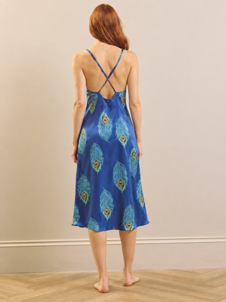 A woman stands barefoot from the back wearing a blue satin slip dress with a peacock feather print and crossed straps at the back, set against a neutral beige wall and light wood floor.