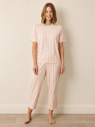 A woman modelling a two-piece pyjama set with a short-sleeve crewneck top and matching cropped pants, both featuring vertical pink and white stripes, set against a neutral beige wall and light wood floor.