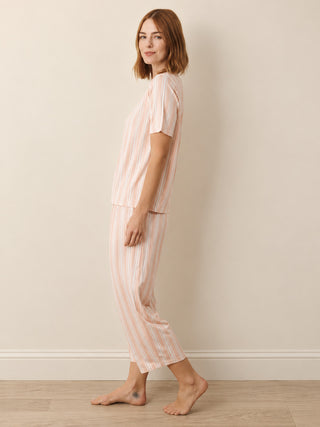 A woman modelling a two-piece pyjama set with short-sleeve top and cropped pants, featuring vertical peach and white stripes, standing barefoot against a neutral beige wall.