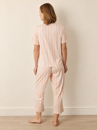 A woman stands with her back to the camera wearing a matching short-sleeve top and cropped pants pyjama set in a pale pink and white vertical stripe pattern, set against a plain beige wall and light wood floor.