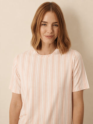A woman with shoulder-length light brown hair wearing a short-sleeve crewneck pyjama top with vertical light pink and white stripes, set against a plain beige background.