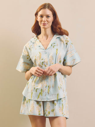 A woman modelling a light blue short-sleeve pyjama set with a green and white wisteria floral print, consisting of a collared button-up top and matching shorts, against a solid beige background.