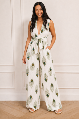 A woman modelling a sleeveless white jumpsuit with a green peacock feather pattern and a matching tie belt at the waist, standing in a room with white panneled walls and light wood flooring.