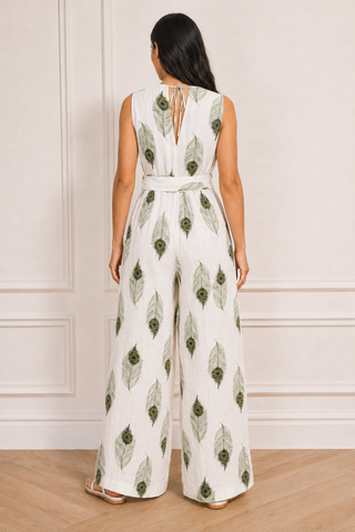 A woman viewed from the back wearing a sleeveless white jumpsuit with an allover green peacock feather print and a matching tie belt at the waist, standing against a cream panneled wall.