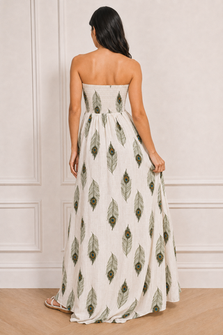 A woman viewed from behind wearing a strapless maxi dress with an off-white base and an all-over green peacock feather print, standing in a room with white panneled walls and light wood flooring.