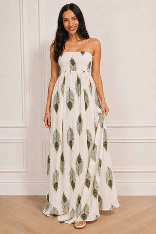 A woman modelling a strapless white maxi dress with a green and brown peacock feather print, standing in front of a white paneled wall.