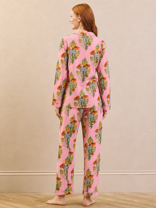 A woman stands barefoot in a studio wearing a pink pyjama set with a colourful floral print, photographed from the back against a beige wall and light wood floor.