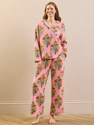 A woman modelling a pink long-sleeve pyjama set with a colourful floral and botanical print, standing against a beige wall and light wood floor.