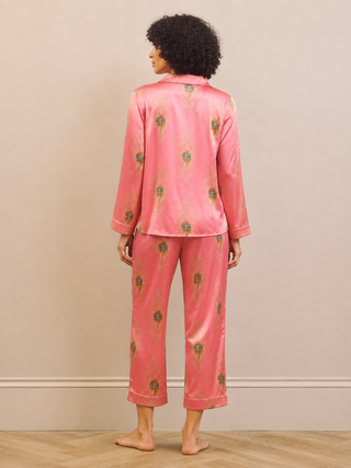 A woman stands barefoot from the back wearing a pink satin pajama set with a small green and gold floral pattern, set against a neutral beige wall and light wood floor.
