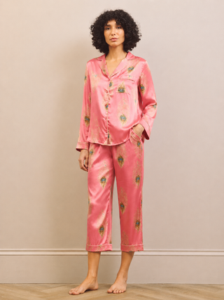 A woman modeling a two-piece satin pajama set in pink with a green and gold floral pattern, standing against a neutral beige wall.