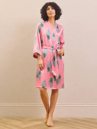 A woman wearing a pink satin dressing gown robe with a blue and green feather print, standing barefoot on a light wood floor against a beige wall.