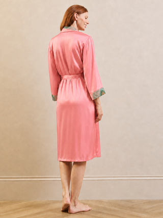 A woman stands barefoot in a studio wearing a pink satin-like robe with green patterned cuffs and collar, photographed from the back against a beige wall and wood floor.