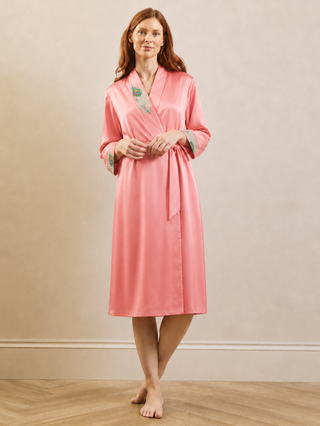 A woman stands barefoot in a studio wearing a pink satin dressing gown robe with embroidered floral details on the collar and cuffs. The background is a warm beige wall with a light wood floor.