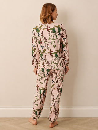 A woman stands barefoot against a plain beige wall, wearing a matching long-sleeve pyjama set with a light pink base and an all-over botanical print of green leaves and dark berries. She is shown from behind, displaying the back of the pajama top and pants.