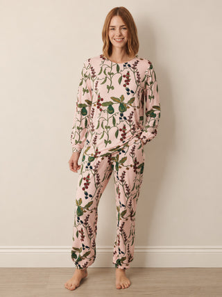 A woman modelling a two-piece long-sleeve pyjama set with a light pink background and a botanical print of green leaves and dark red berries, standing against a neutral beige wall.