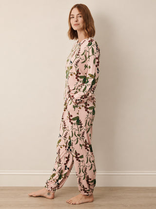 A woman stands barefoot in profile wearing a long-sleeve, loose-fitting pyjama set with a pink base and an allover botanical print in green and dark brown, set against a neutral beige wall and light wood floor.