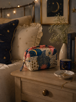 Colourful patchwork cosmetic bag on a wooden nightstand with decorative pillows and books in the background.