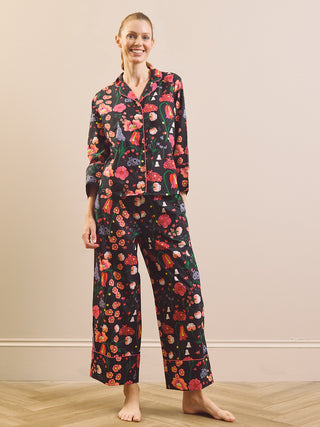 A woman stands barefoot in a bedroom setting wearing a matching pyjama set with a dark background and a colourful floral print. The set includes a long-sleeve button-up top and wide-leg pants.