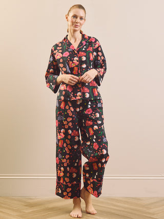 A woman stands barefoot in a bedroom setting wearing a matching pyjama set with a dark background and a colourful fruit print. The set includes a long-sleeve button-up shirt and wide-leg pants.