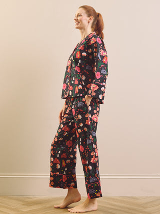 A woman wearing a two-piece floral pyjama set with a long-sleeve button-up top and matching pants, standing in profile against a beige wall.