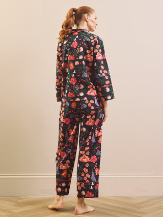 A woman stands barefoot in a room wearing a matching two-piece pyjama set with a dark background and a dense floral print in red, pink, orange, and purple. The set includes a long-sleeve top and cropped pants.